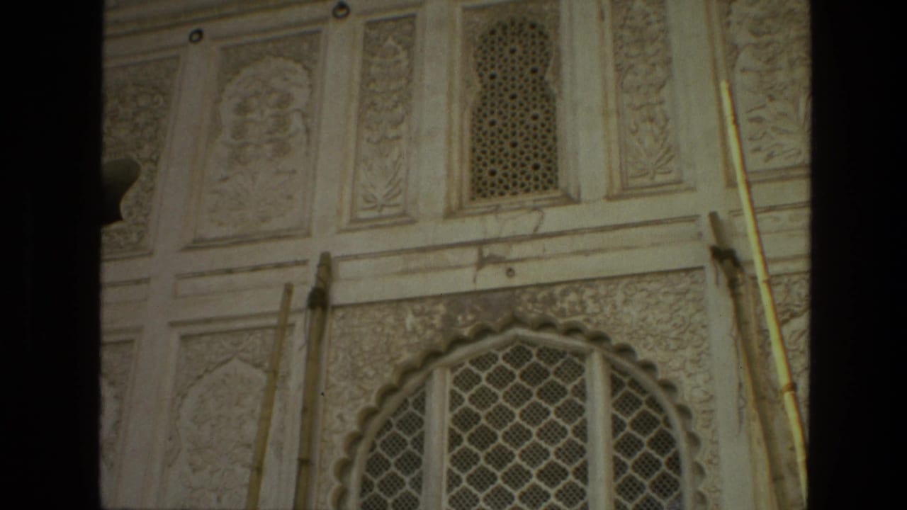 1974 India home-movie frame of an ornate pale temple facade with carved panels, small lattice openings, vertical rods, and a scalloped arch below.