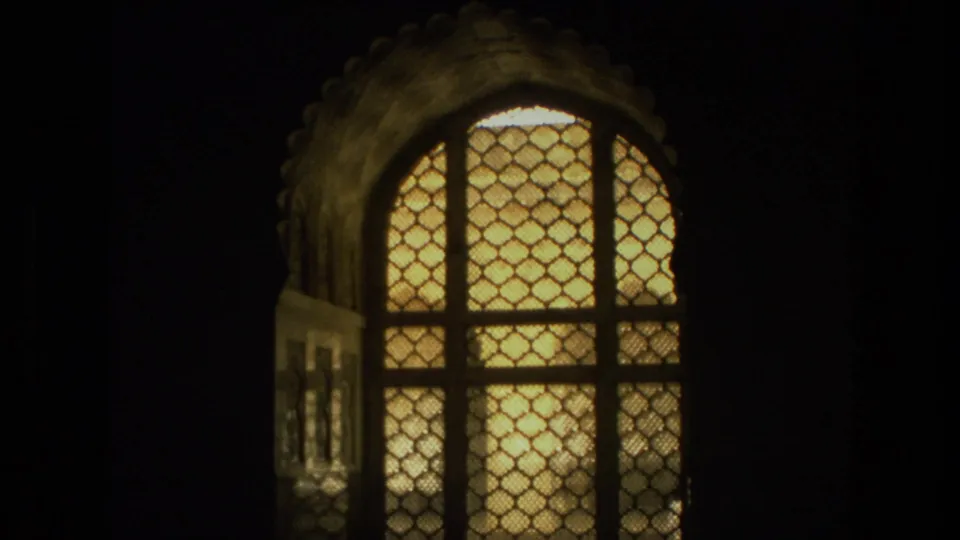 1974 India home-movie frame showing a glowing arched lattice window, scalloped surround, dark foreground framing, and patterned screen detail.