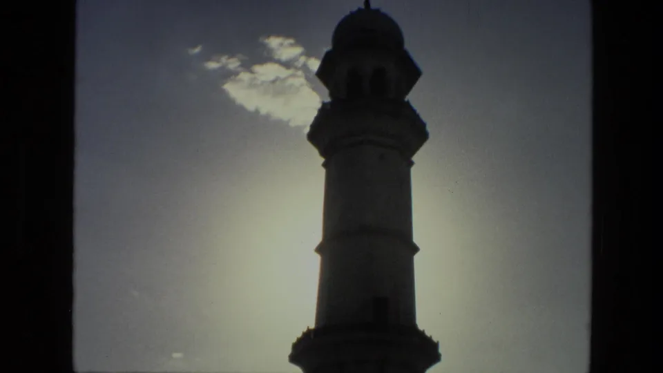 1974 India 8mm view of a minaret-like tower in silhouette against bright sky and scattered cloud.