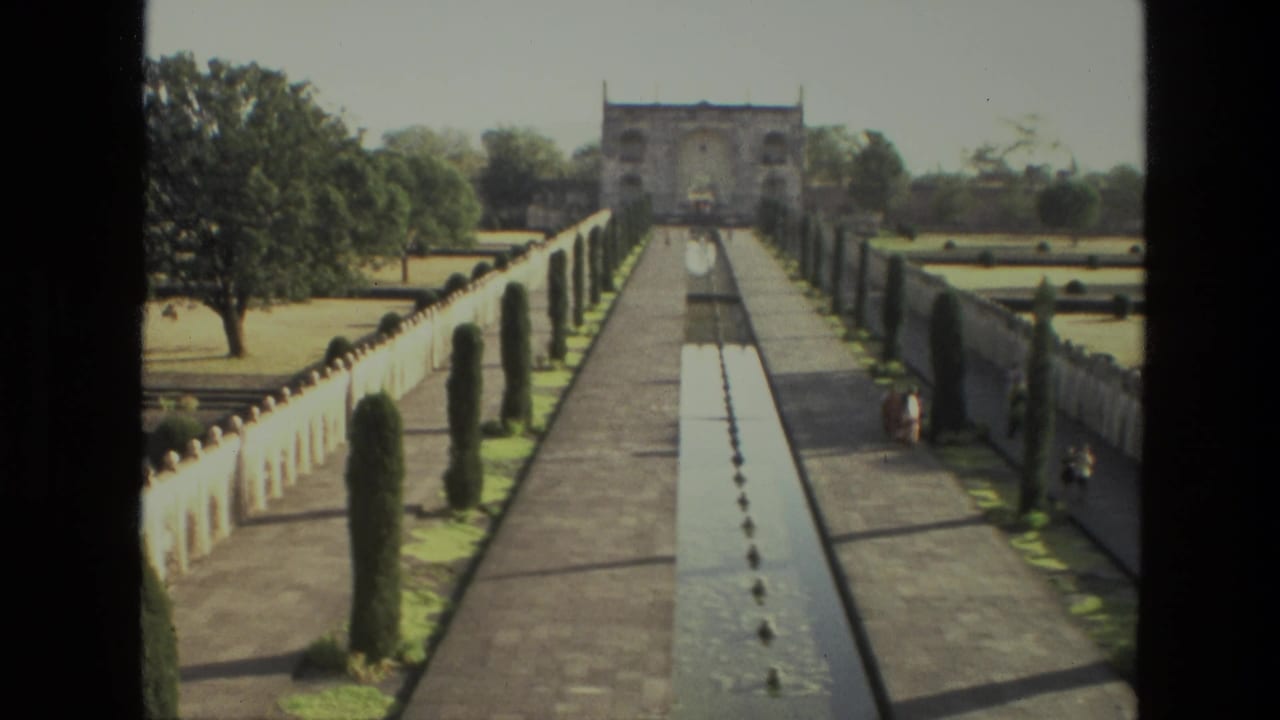 1974 India home-movie view of a long garden walkway with a central pool, clipped trees, low walls, and a distant gateway.