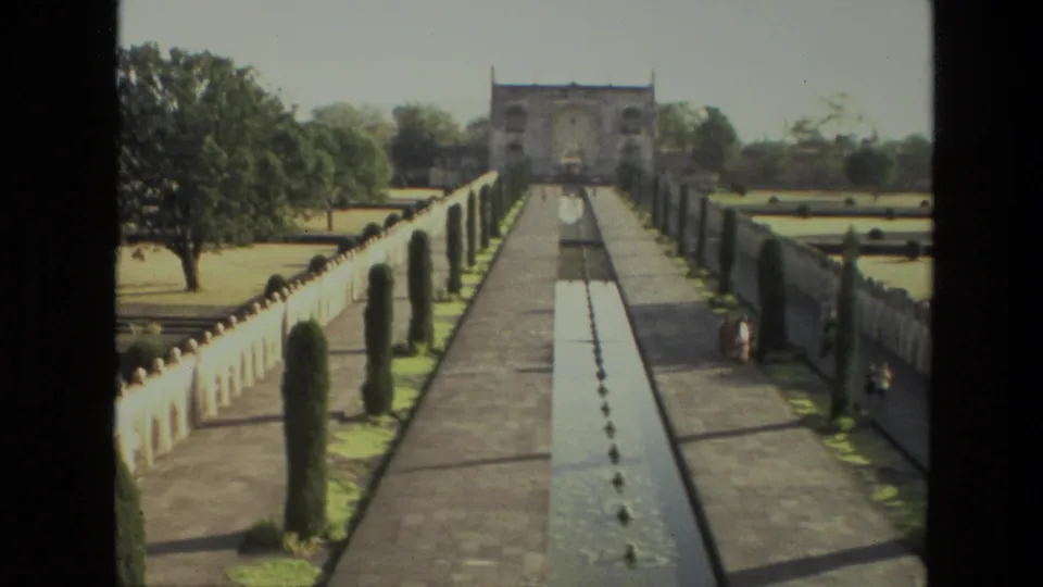 1974 India home-movie view of a long garden walkway with a central pool, clipped trees, low walls, and a distant gateway.