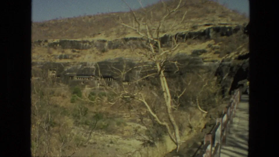 1974 India 8mm frame showing a dry hillside landscape with leafless trees, rock faces, and cave-like openings cut into the slope.