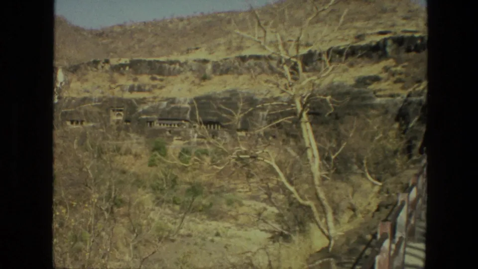 1974 India home-movie frame of a rocky hillside with cave-like structures, dry vegetation, bare trees, and a walkway along the right edge.