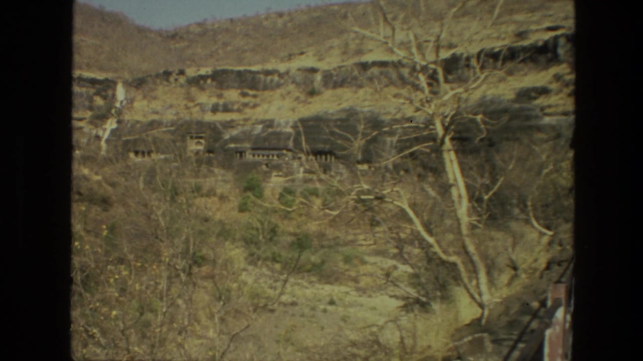 Dry hillside with sparse trees and distant rock-cut openings framed by dark film borders.