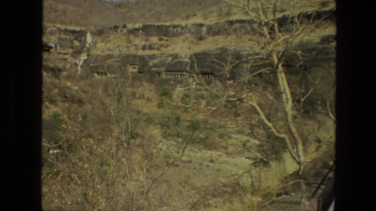 1974 India frame showing a rugged hillside with possible rock-cut caves behind dry scrub and bare trees.