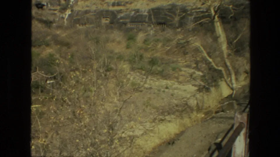 Motion-softened 8mm frame from India in 1974 with dry vegetation and distant openings along a rock face.