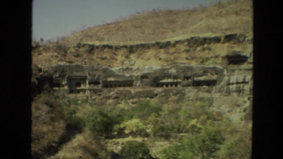 A distant 1974 India view shows cave-like temple facades set into a dry hillside with scattered vegetation.