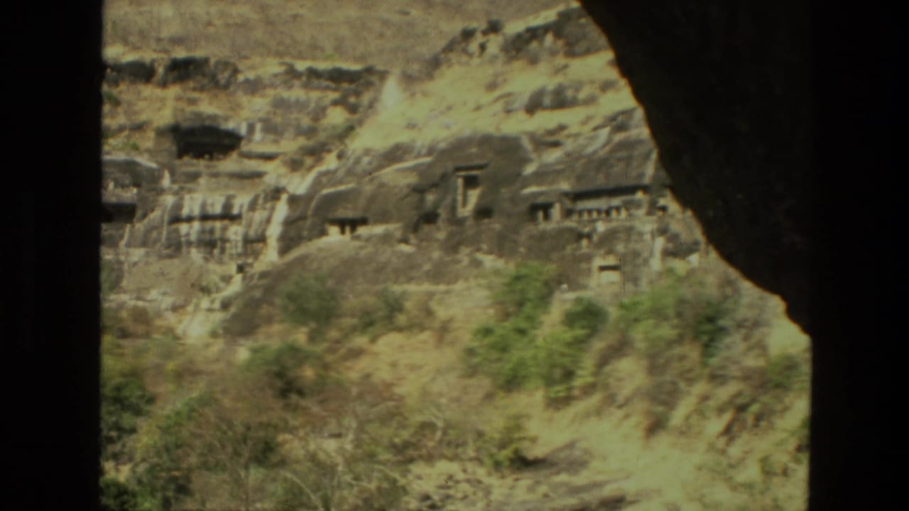 1974 India travel-film frame of cave entrances and carved stonework across a rugged slope, partly obscured by shadowed foreground.