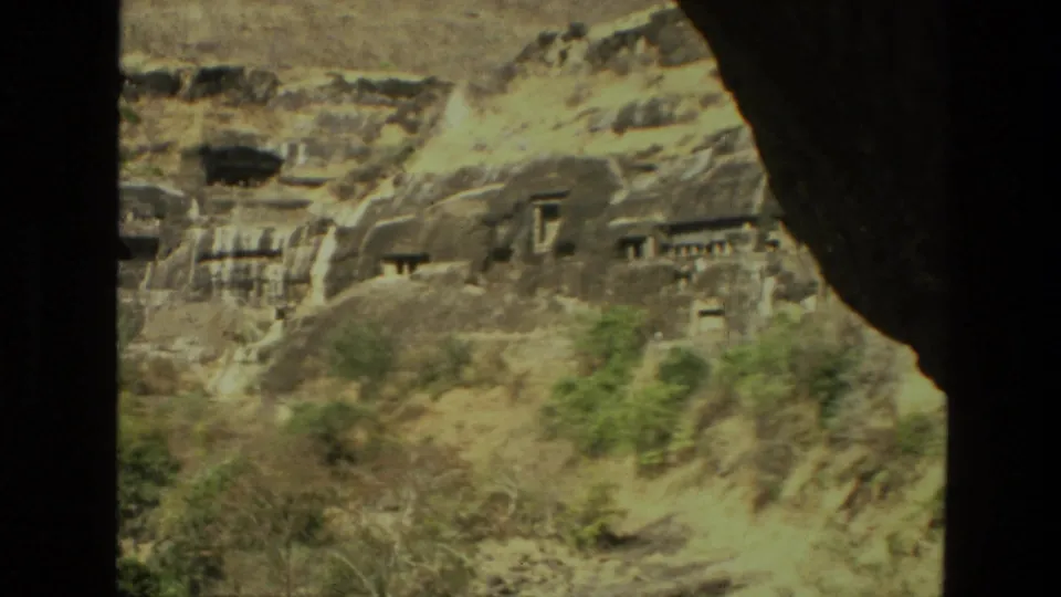 1974 India travel-film frame of cave entrances and carved stonework across a rugged slope, partly obscured by shadowed foreground.