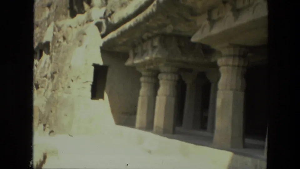 1974 India travel frame of a pillared rock-cut cave entrance, with bright sunlit foreground and dark interior shadows.