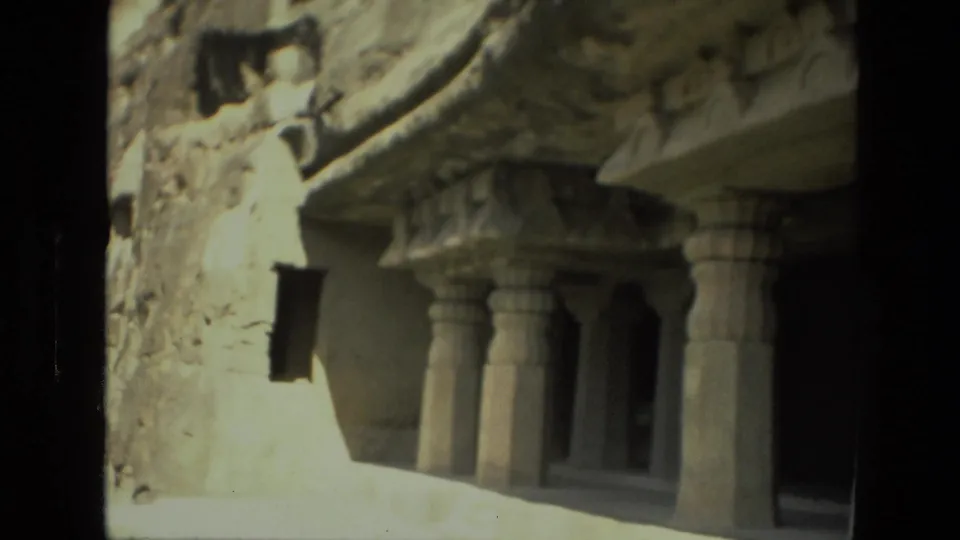 1974 8mm view in India of a rock-cut temple facade with carved pillars, shadowed openings, and weathered stone surfaces.