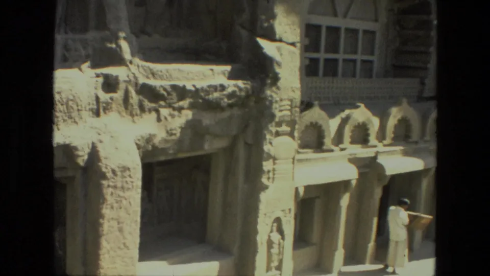 1974 8mm view of an Indian rock-cut temple facade with arched stone details, pillars, reliefs, and a distant visitor for scale.
