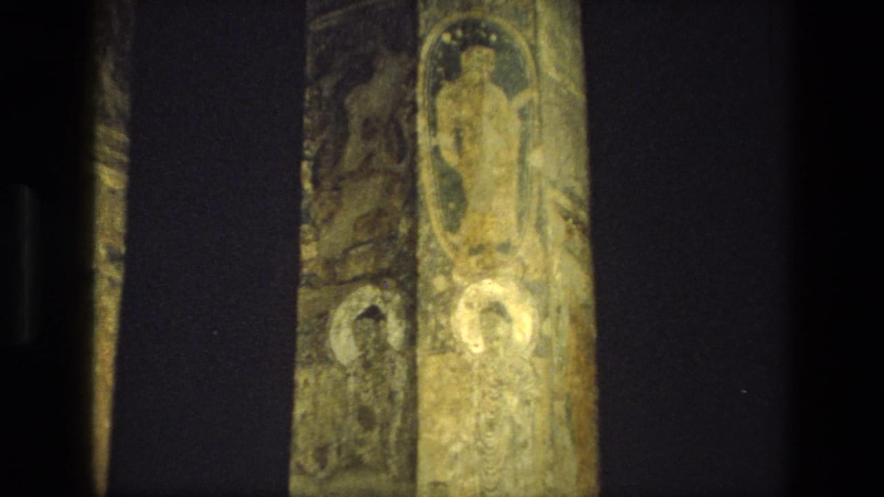 Dim cave-temple interior with painted vertical stone pillars and shadowed mural surfaces.