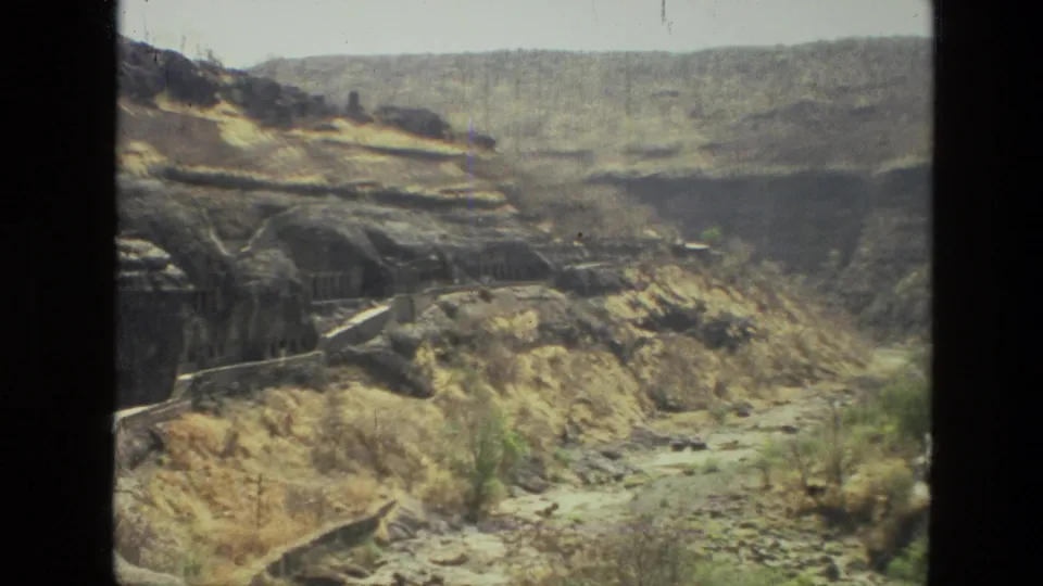 1974 India 8mm frame widening to show rock-cut structures set into a rugged hillside with dry vegetation and distant mountain ridges.