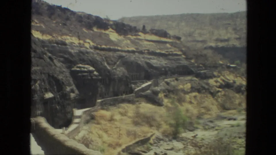 1974 India 8mm frame showing the rock-cut hillside from a visitor vantage, with a path, dry grasses, and layered terrain in the distance.