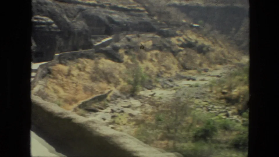 Handheld 8mm frame from India in 1974 showing cave openings, canyon slopes, shrubs and a rocky streambed below.