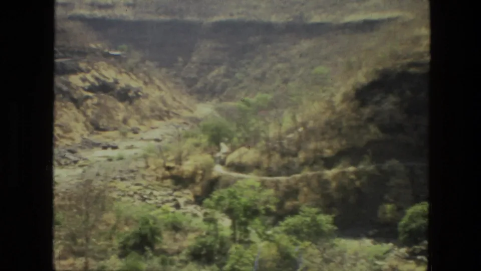 A 1974 India home-movie frame looks across the rocky canyon floor toward layered cliffs and scattered green shrubs.