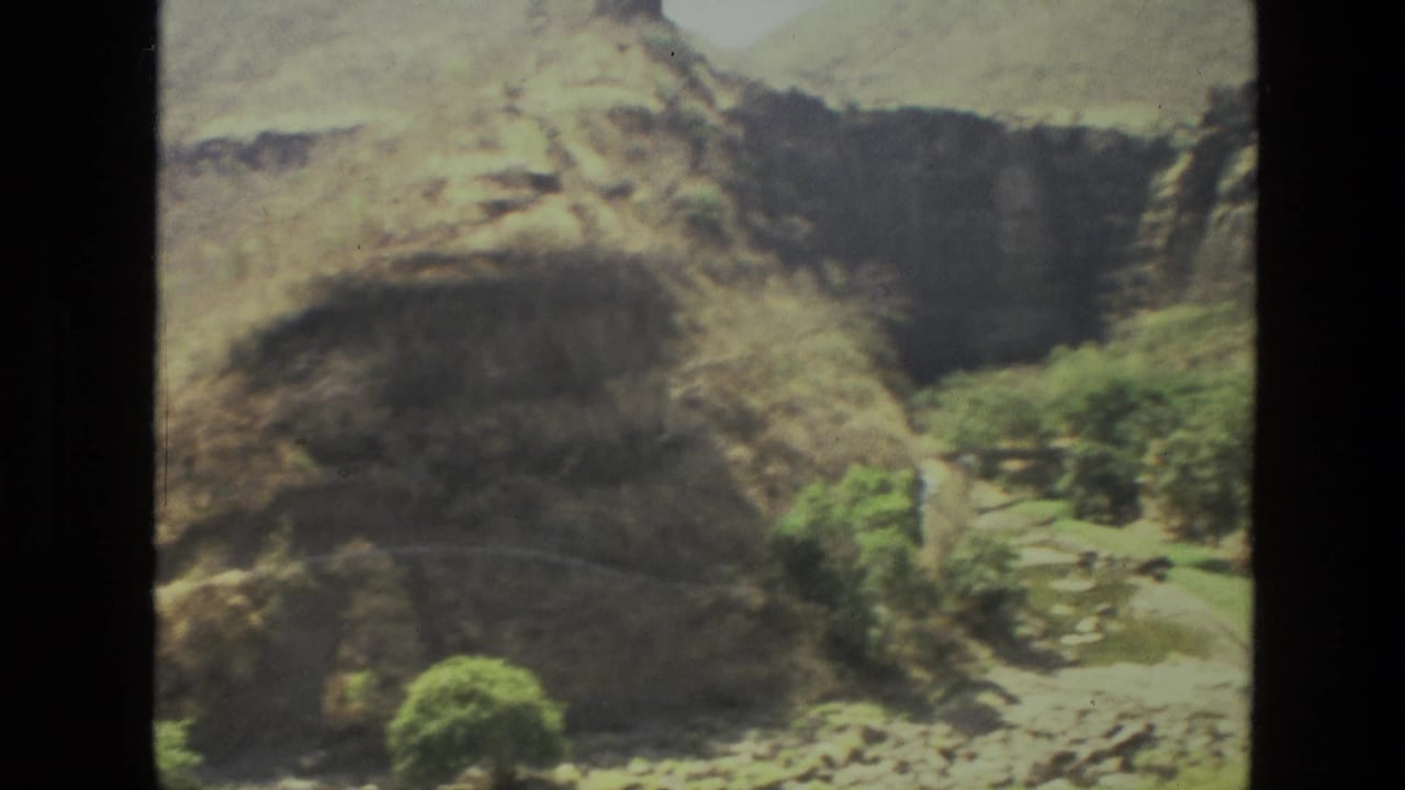 1974 India home-movie frame showing steep rock walls, scrubby trees, and a narrow ravine running through the hills.
