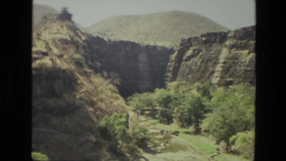 1974 8mm frame from India with layered cliffs, a rounded distant hill, and green trees following the ravine below.