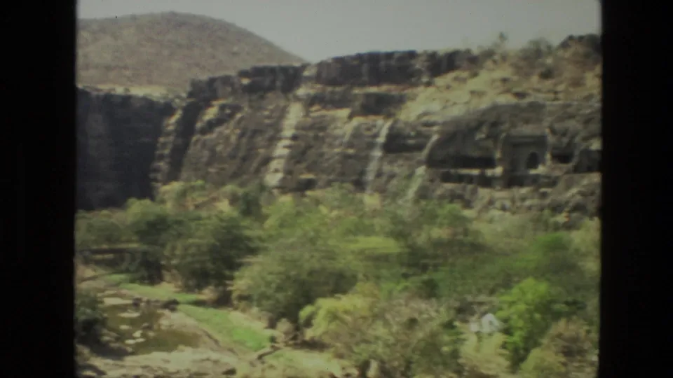 1974 India travel frame showing a cliffside with scrub vegetation, a winding path, and a dark rock recess or shadowed opening.