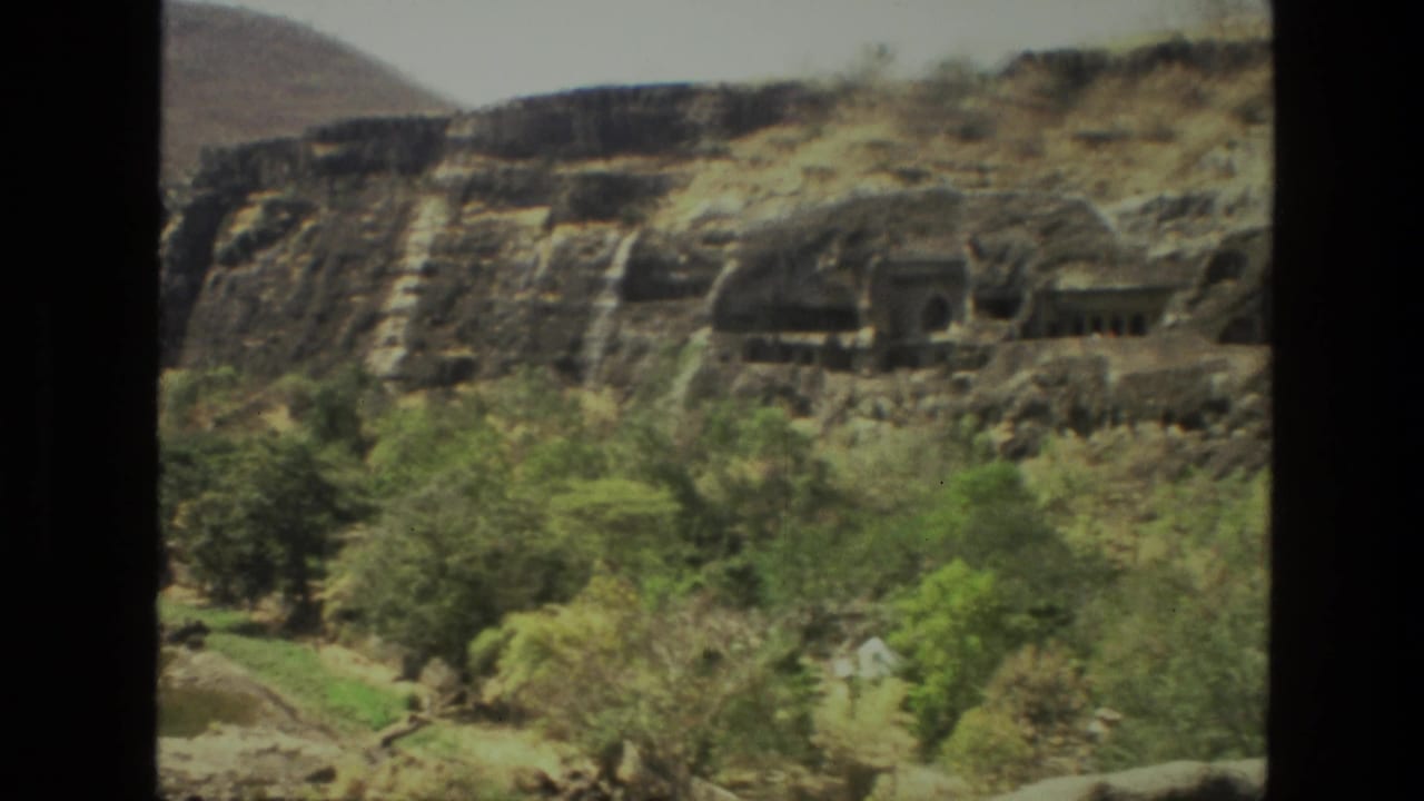 Rock-cut temple entrances across a dry cliff with trees below.