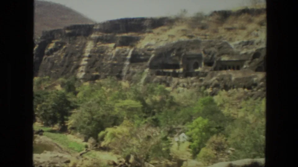 1974 8mm view in India of cave-like temple openings carved into a rocky hillside above green trees.