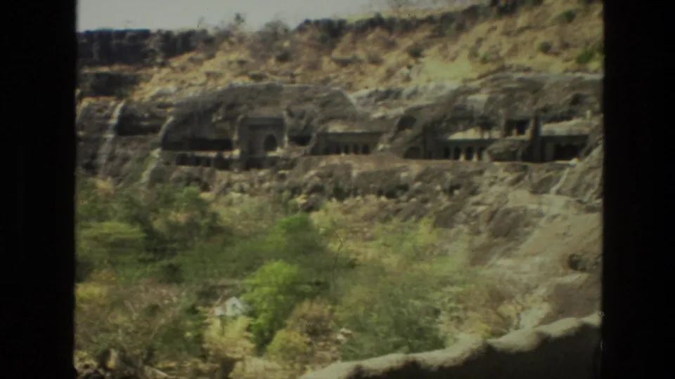 A soft 8mm frame shows Indian rock-cut temple openings across a rugged cliff, with vegetation on the lower slope.