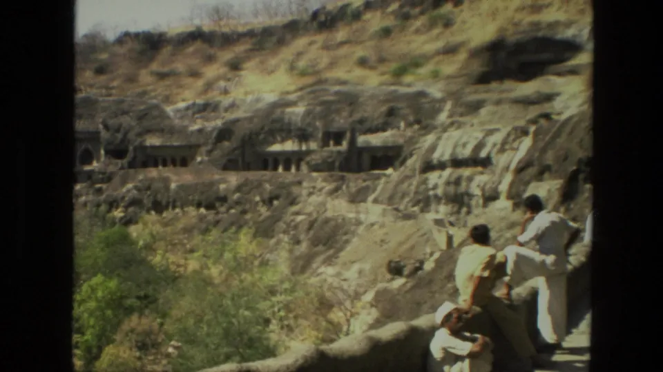 Visitors at a lookout watch the rock-cut temple complex across the rocky slope in this 1974 India travel frame.