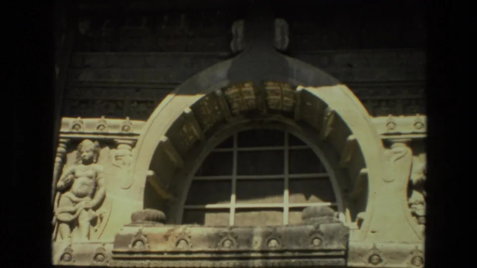 Close 8mm view of an ornate carved stone facade in India, showing an arched opening and sculptural figures.