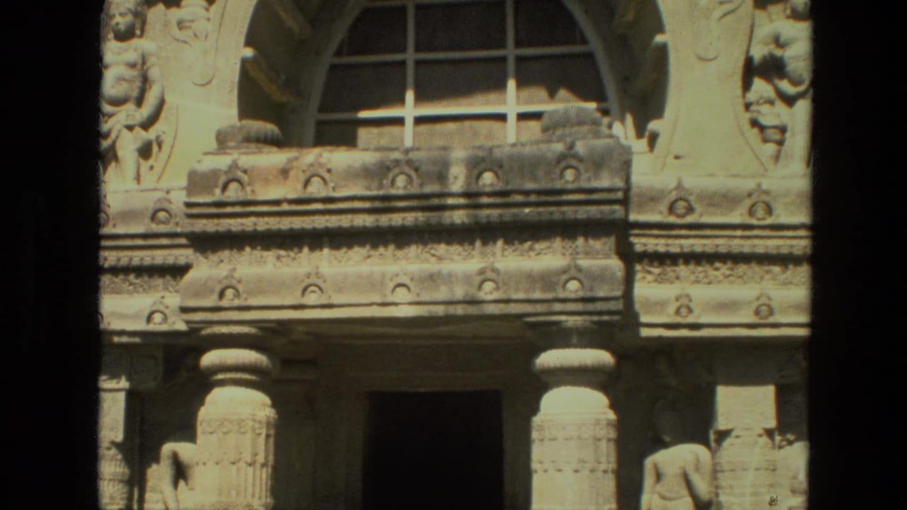 Sunlit carved stone facade with arched window, pillars, relief bands, and a dark doorway.