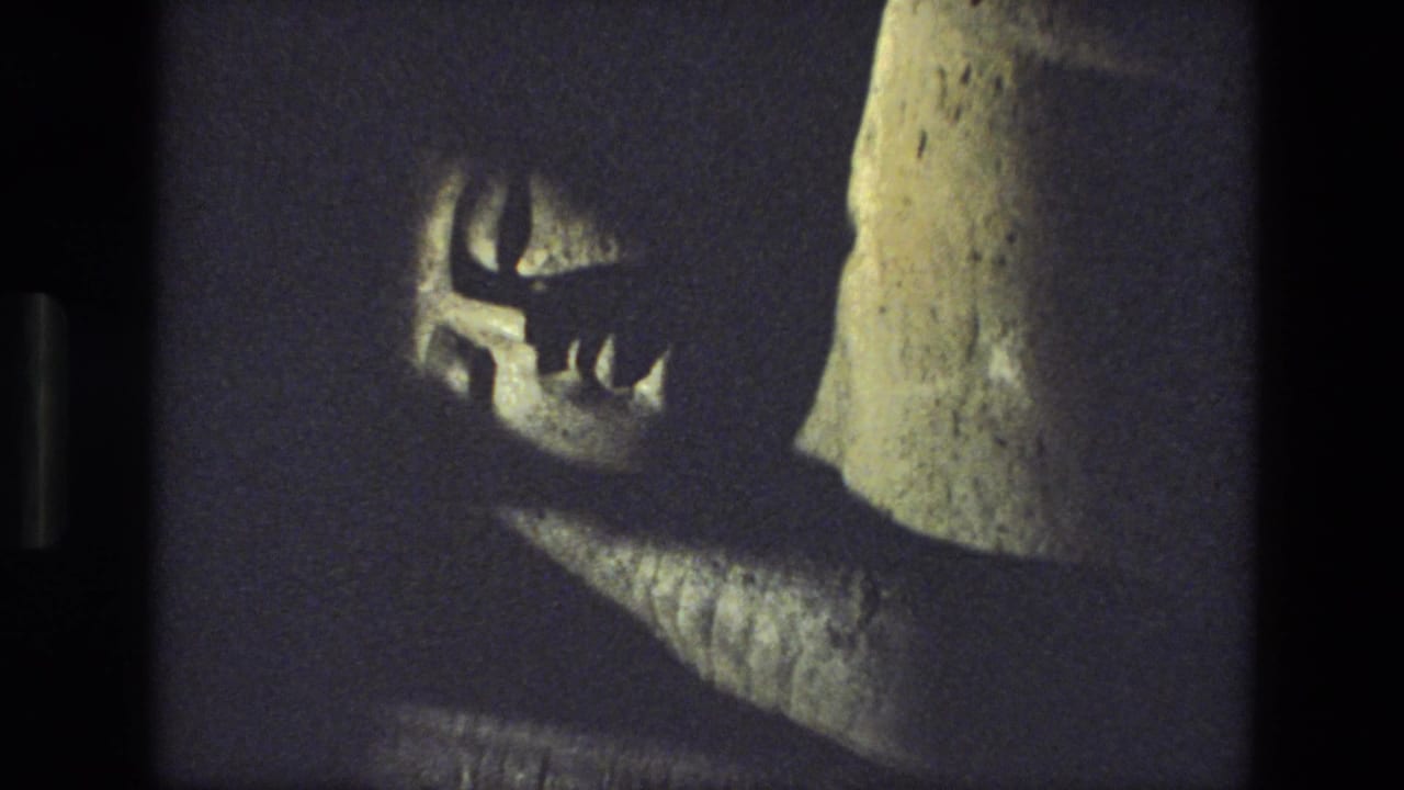 Dim 8mm frame showing part of a reclining stone statue with a carved face and shadowed support.