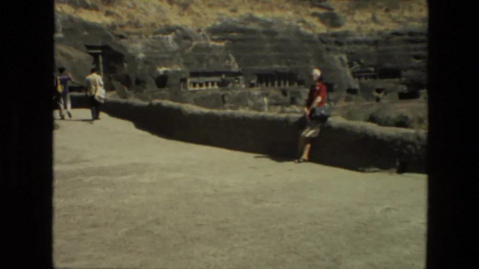 1974 8mm India travel frame showing a woman in a red shirt and white hat near a low wall, with rock-cut cave structures behind her.