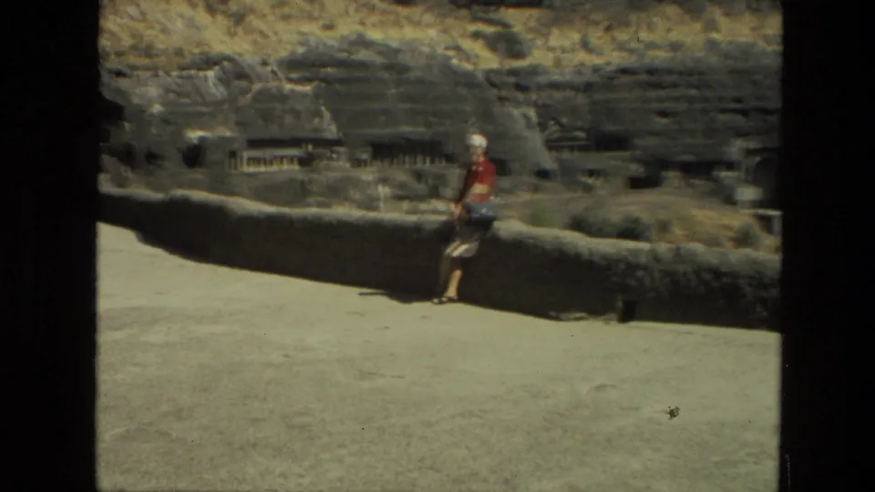 1974 8mm India travel frame with a woman in a red shirt and white hat leaning near a low wall before carved rock and cave openings.