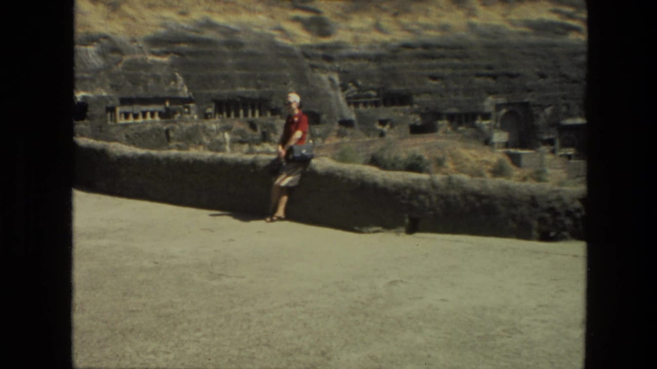 Woman in red stands by a low stone wall with rock-cut cave openings behind her.