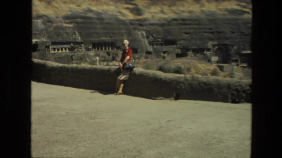1974 India 8mm frame showing a woman in red and a white head covering beside a wall overlooking rock-cut cave architecture.