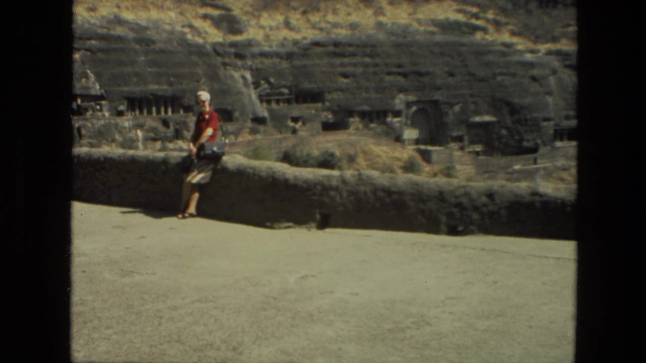 Home-movie frame from India in 1974 with a woman at the left of a stone overlook and rock-cut cave features across the background.