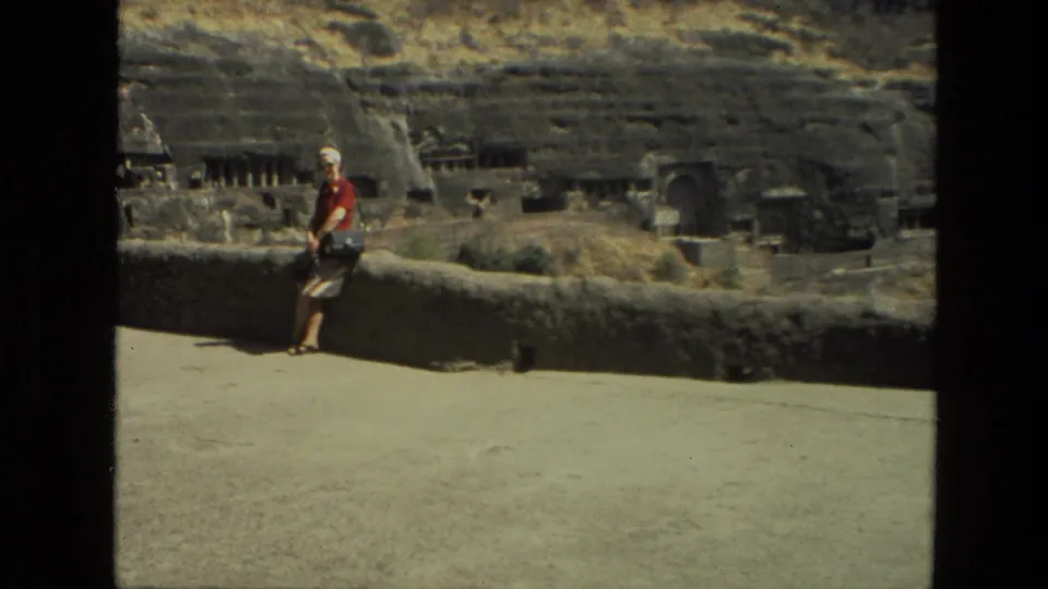 Home-movie frame from India in 1974 with a woman at the left of a stone overlook and rock-cut cave features across the background.