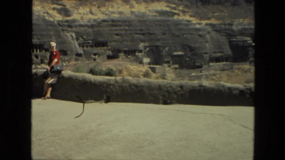 1974 India travel footage frame with the visitor near the frame edge and the pan emphasizing the rock-cut cave structures and wall.