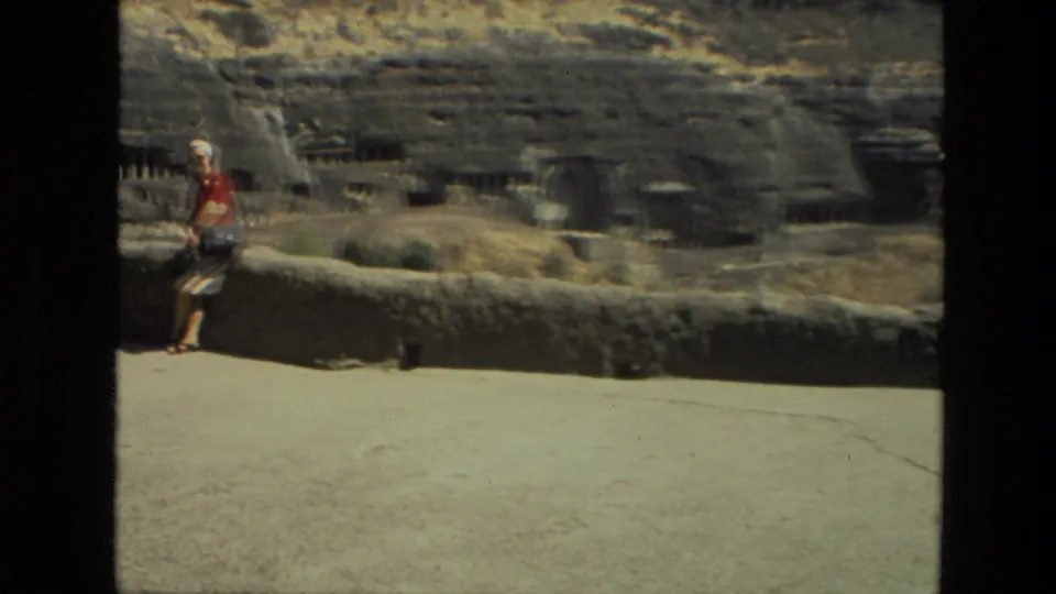Final 1974 India 8mm frame of a woman beside a stone wall as the pan settles on cave openings and carved hillside architecture.