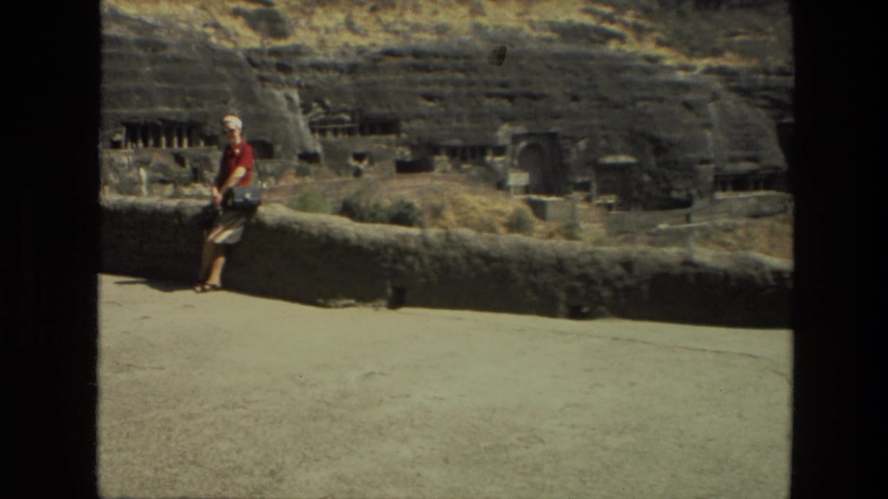 Tourist in a red shirt leaning on a low stone wall with rock-cut ruins behind.