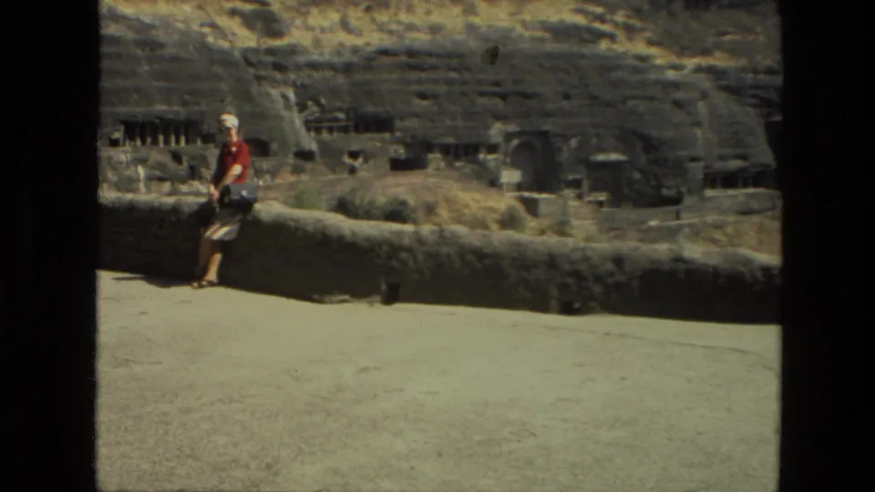 1974 India travel footage frame showing a tourist in a red shirt and white hat leaning at a stone wall before rock-cut ruins.