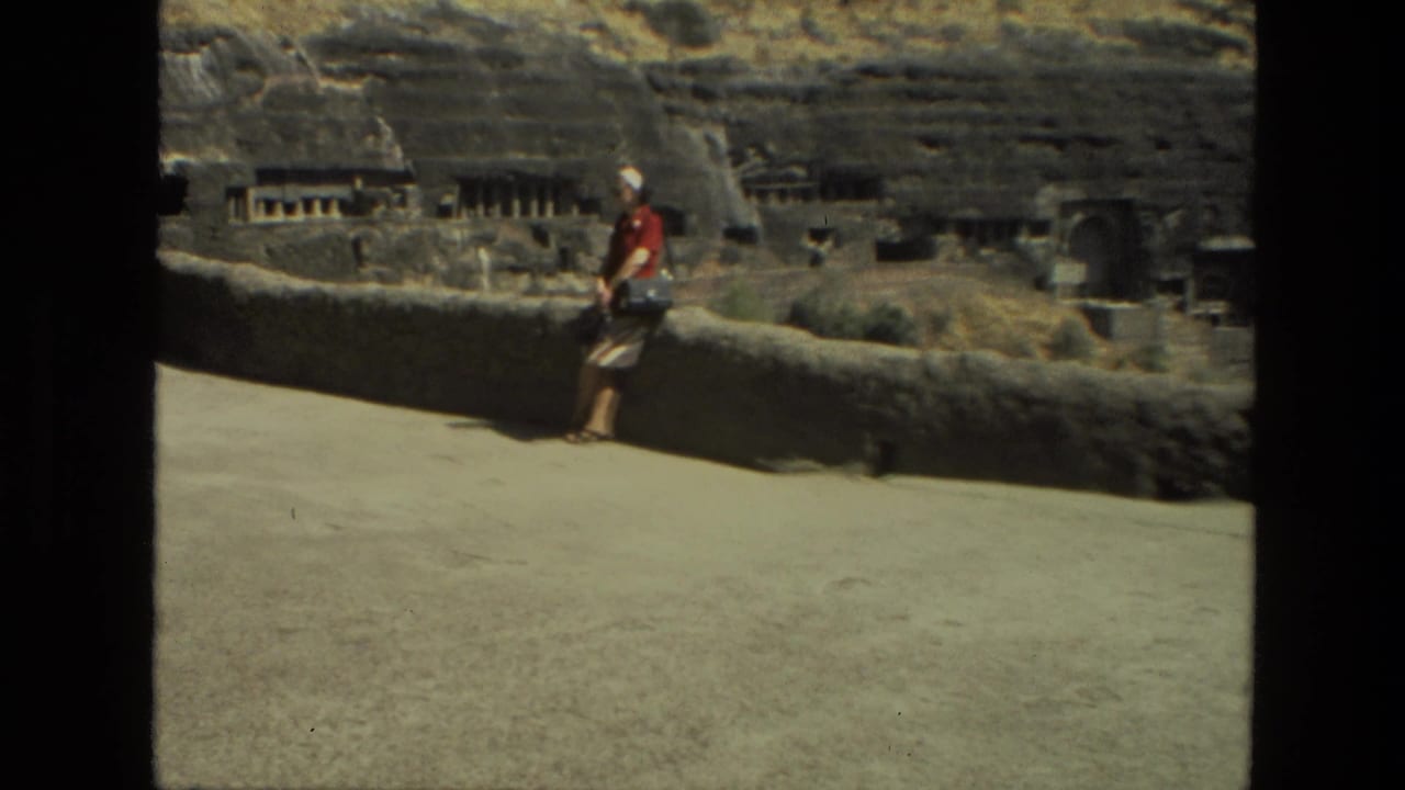 A sunlit 1974 India frame shows a visitor near a low wall, with carved stone openings and hillside ruins behind.