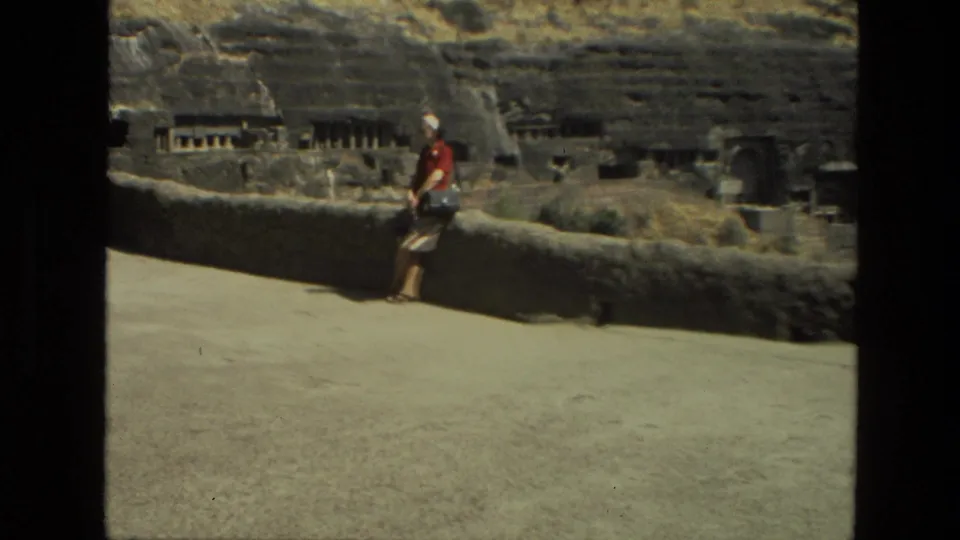 A sunlit 1974 India frame shows a visitor near a low wall, with carved stone openings and hillside ruins behind.
