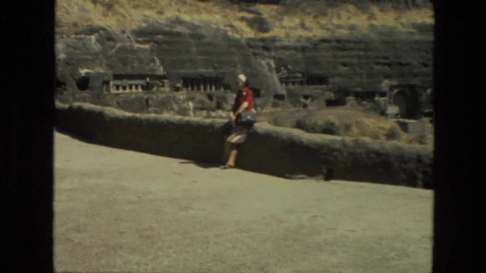 Home-movie frame from 1974 India showing a tourist at a low wall before weathered rock-cut structures.