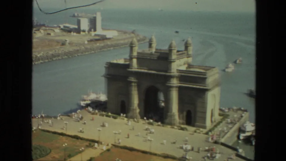Soft 8mm frame of the Gateway of India in Mumbai, showing the plaza, harbor channel, small boats, and distant waterfront structures.