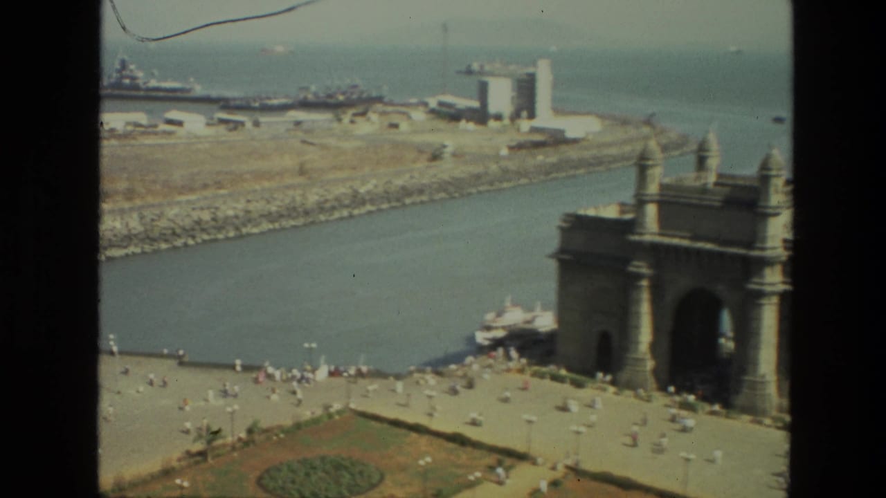 1974 India home-movie view of the Gateway of India waterfront, with harbor boats, a breakwater, distant buildings, and visitors below.