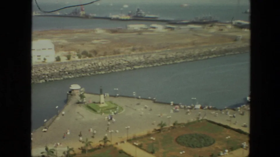 1974 India waterfront view showing a circular monument on the promenade, harbor channel, breakwater, distant buildings, and tiny visitor figures.
