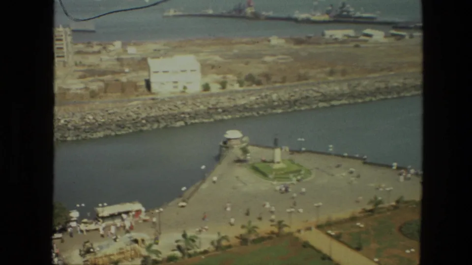1974 India harbor scene with a waterfront monument, promenade visitors, boats, breakwater, and buildings across the water.