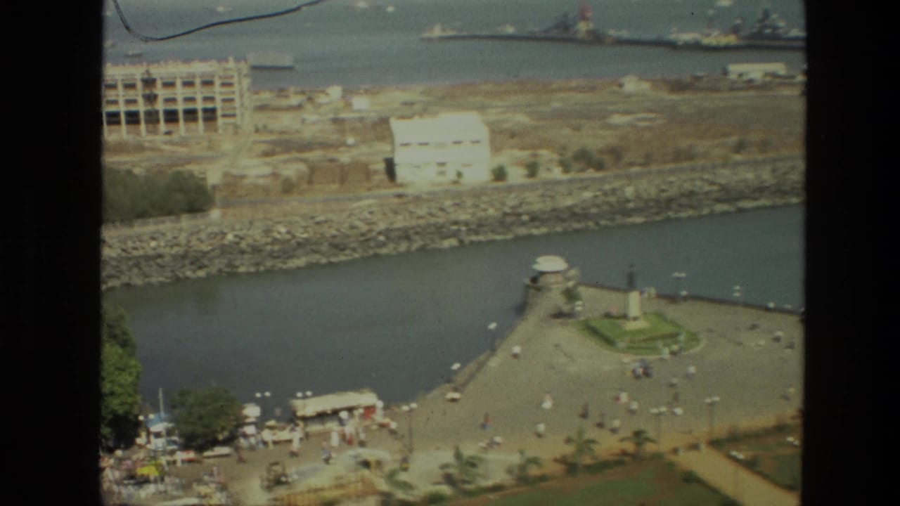 1974 India home-movie frame showing a high-angle waterfront with a promenade, central monument, distant ships, and shoreline construction.