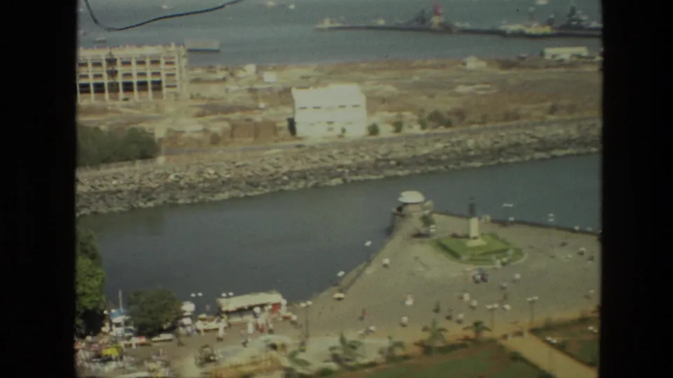 1974 India home-movie frame showing a high-angle waterfront with a promenade, central monument, distant ships, and shoreline construction.